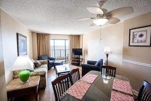 A hotel room with a dining table, chairs, a sofa, a TV, a ceiling fan, and a window with curtains.