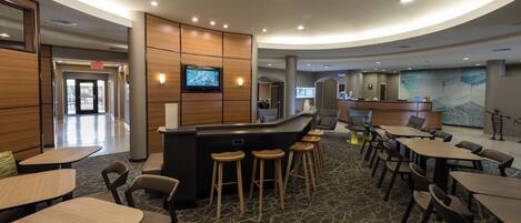 A modern hotel lobby with a curved reception desk, bar stools, and a seating area with tables and chairs.