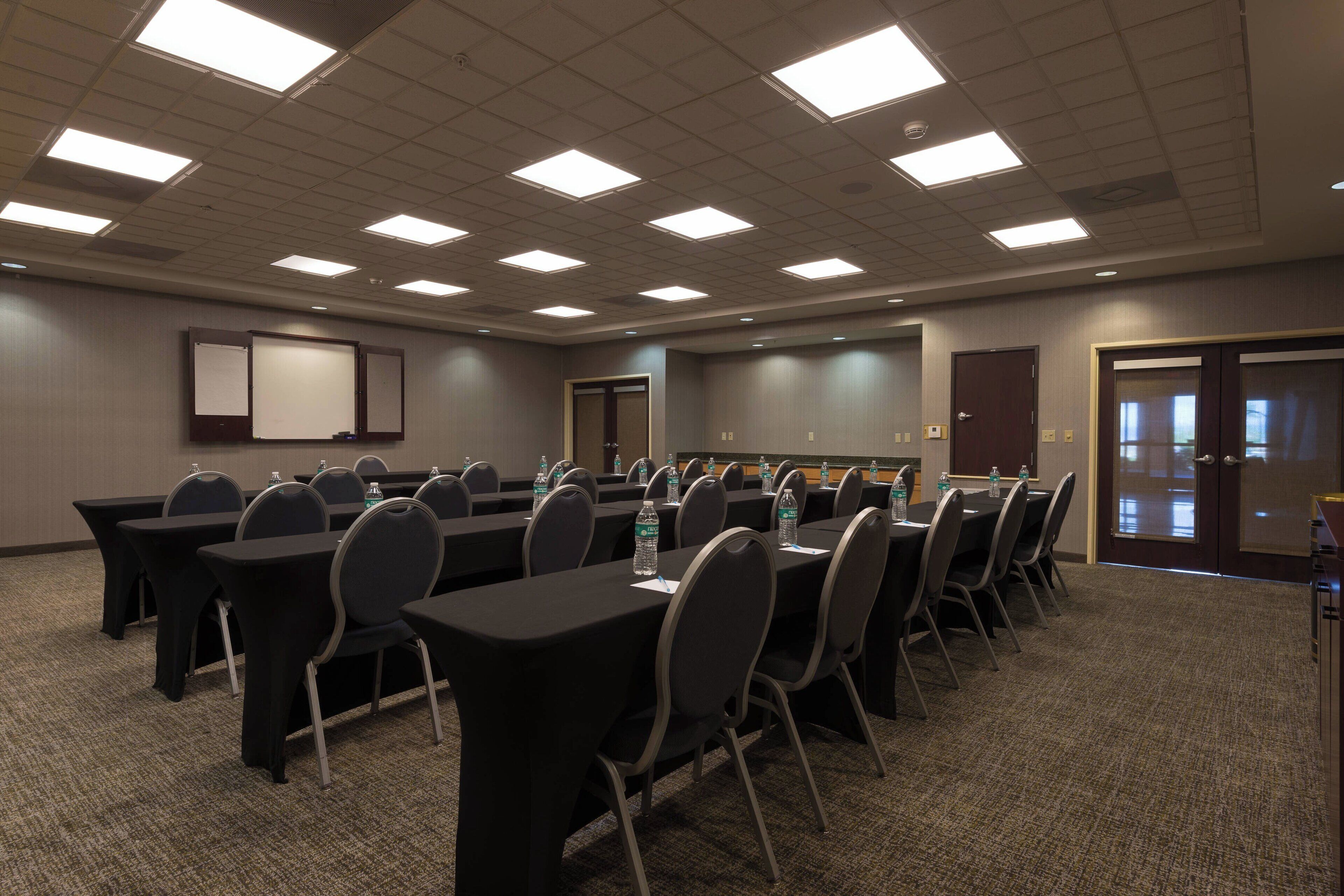 A conference room with multiple tables and chairs, equipped with water bottles, and a projection screen on the wall.
