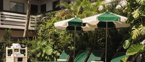 A pool area with lounge chairs and umbrellas, surrounded by lush greenery and a building in the background.