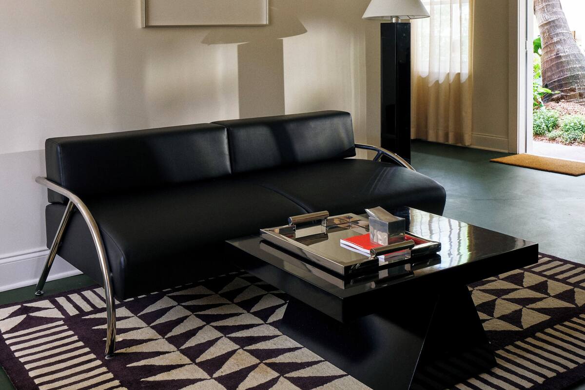 Una sala de estar moderna con un sofá de cuero negro, una mesa de centro de vidrio, una alfombra estampada, un cuadro en la pared y una lámpara.