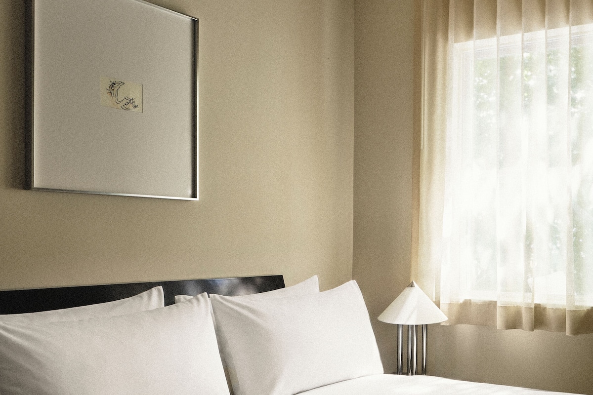Una habitación de hotel con cama, lámpara de noche, un cuadro en la pared y una ventana con cortinas.