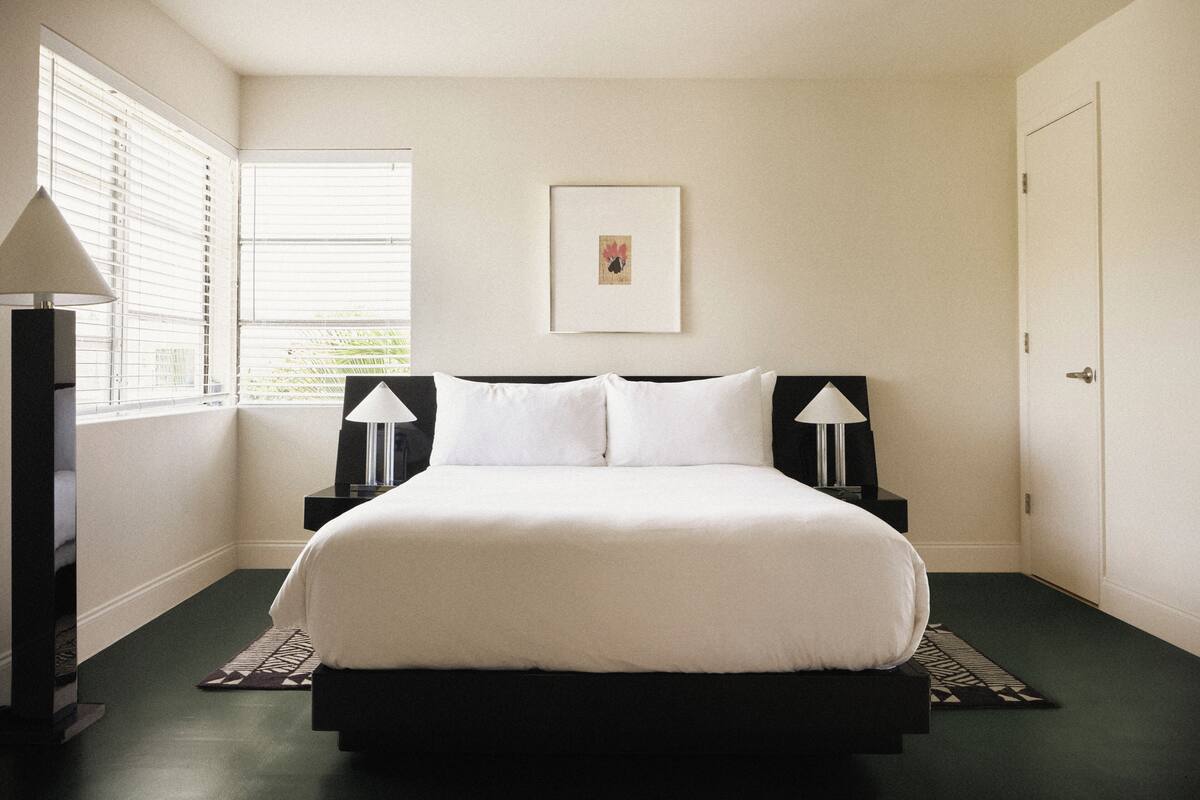 Un dormitorio con cama, dos lámparas de noche, un cuadro en la pared y una ventana con persianas.
