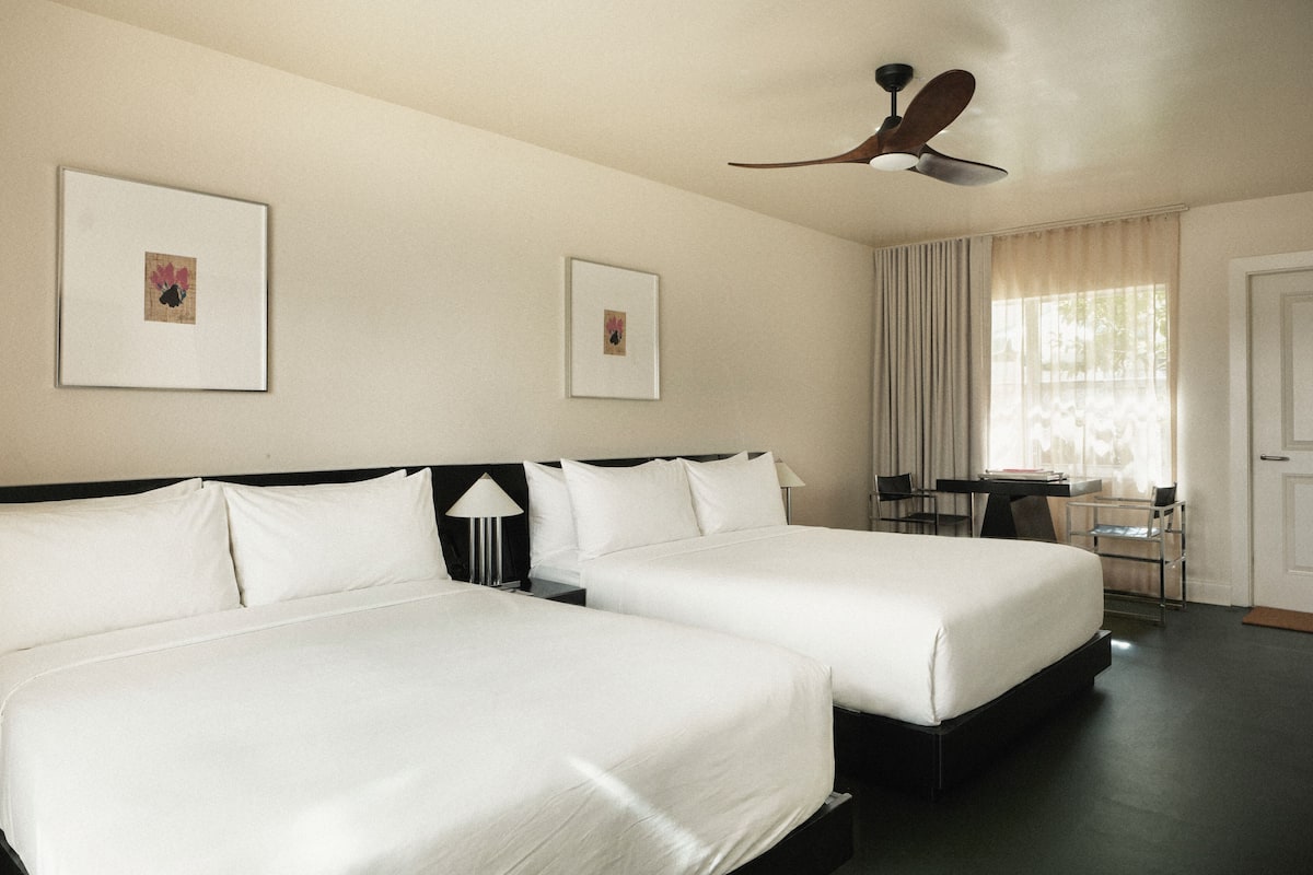 Habitación de hotel con dos camas, un escritorio, una silla, un ventilador de techo y dos cuadros en la pared.