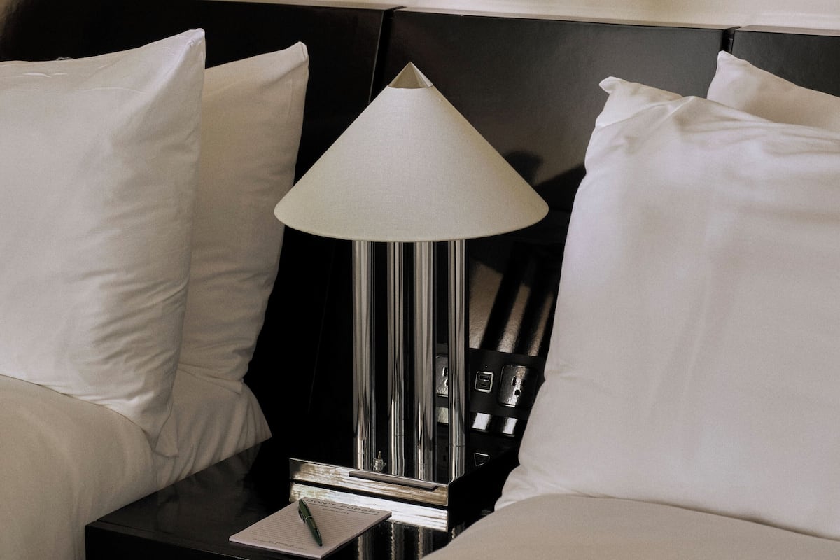 Una cama bien hecha con sábanas blancas, una mesita de noche negra con lámpara y un cuadro colgado en la pared.