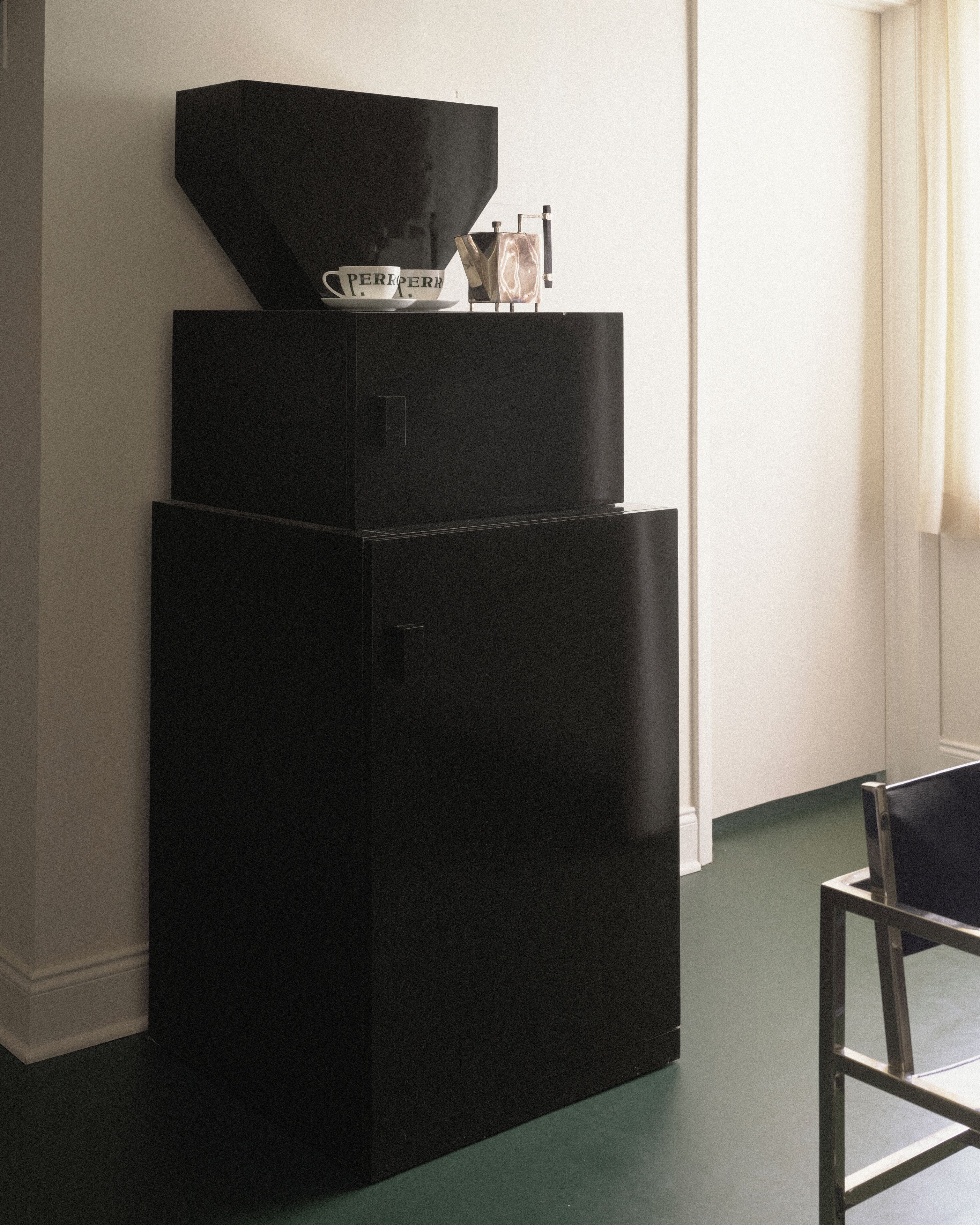 A black refrigerator with a coffee maker and a cup on top, a chair, and a window with curtains.