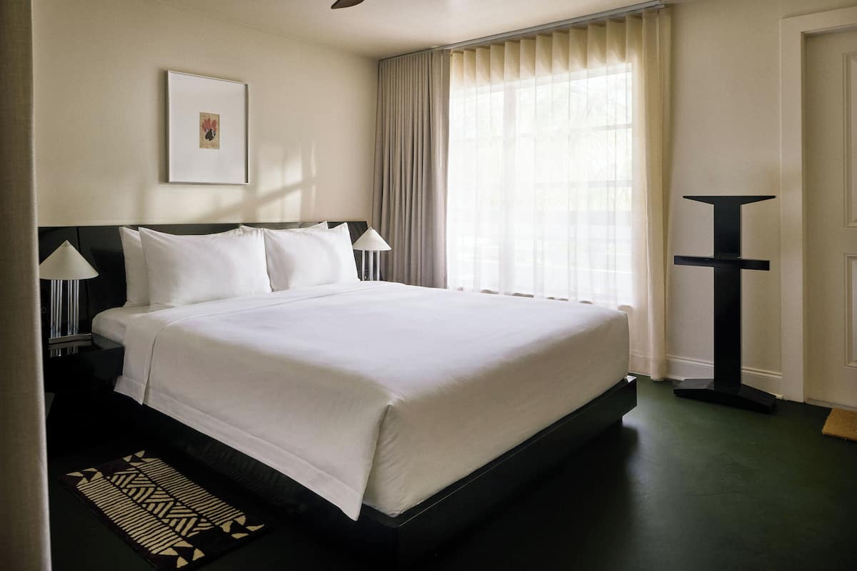 Habitación de hotel con una cama grande, una mesita de noche con lámpara, un cuadro en la pared y una ventana con cortinas.