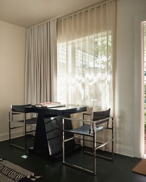 Ein Zimmer mit Tisch und zwei Stühlen, einem Fenster mit durchscheinenden Vorhängen und Blick ins Grüne.