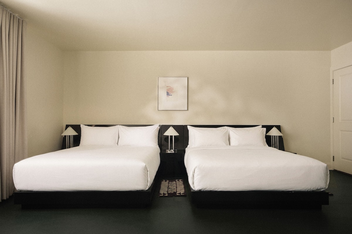 Una habitación de hotel con dos camas, una mesita de noche, una lámpara y un cuadro en la pared.