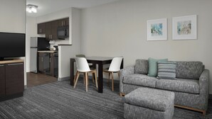 Un salon moderne avec un canapé gris, un téléviseur, une table à manger avec des chaises, et un coin cuisine à l’arrière-plan.