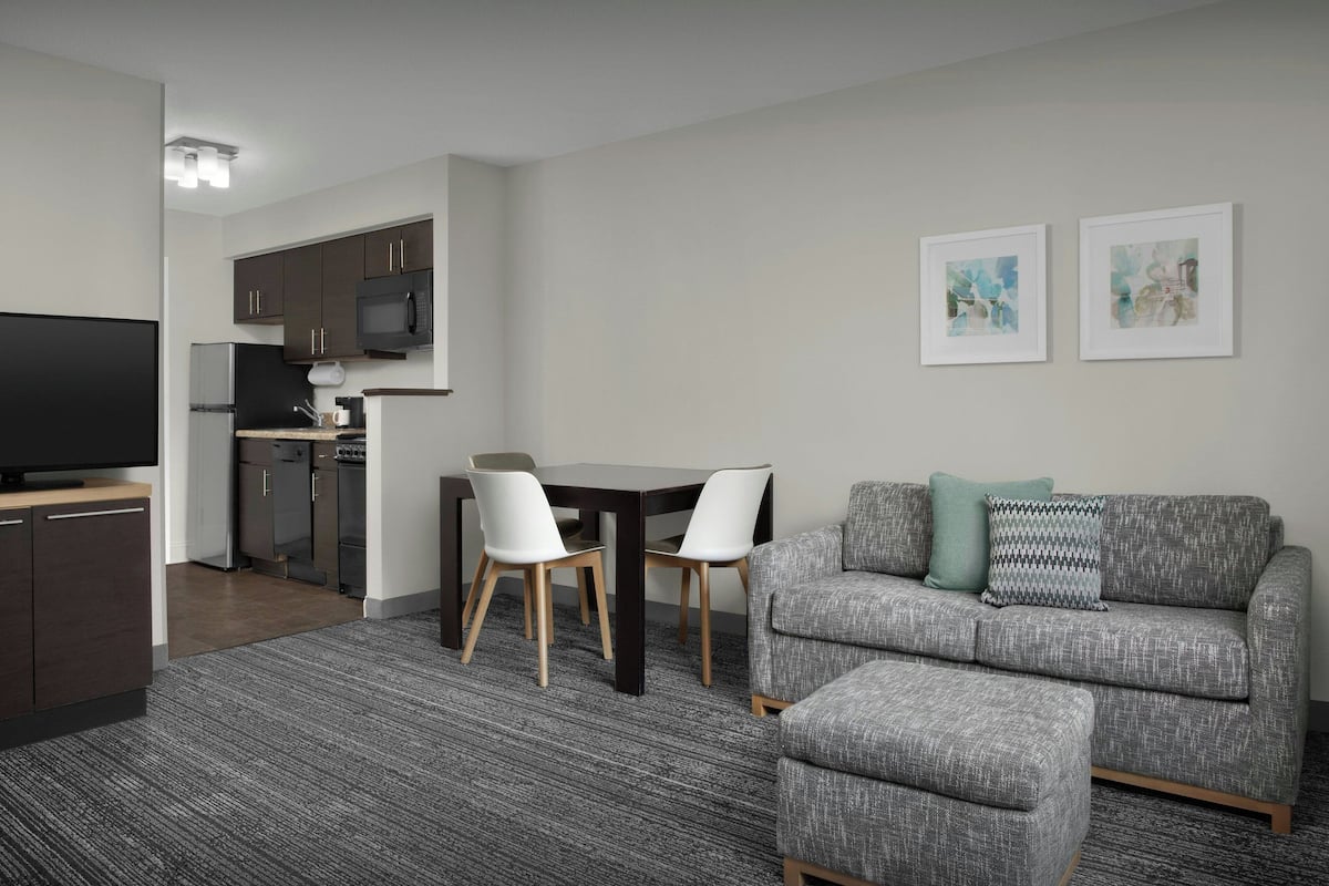 Ein modernes Wohnzimmer mit einem grauen Sofa, einem Fernseher, einem Esstisch mit Stühlen und einer Küche im Hintergrund.