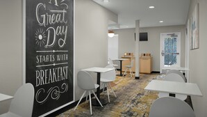 Un espace moderne pour le petit-déjeuner, avec des tables et des chaises blanches, un tableau noir où sont inscrites des phrases motivantes, et une cuisine en arrière-plan.