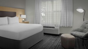Une chambre d’hôtel avec un grand lit, une table de chevet avec une lampe, une chaise et une fenêtre avec des rideaux légers.