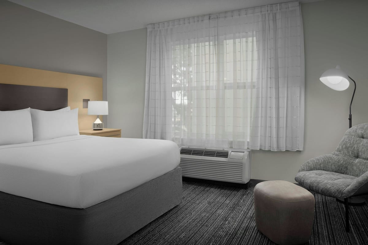 Ein Hotelzimmer mit einem großen Bett, einem Nachttisch mit Lampe, einem Sessel und einem Fenster mit durchsichtigen Vorhängen.