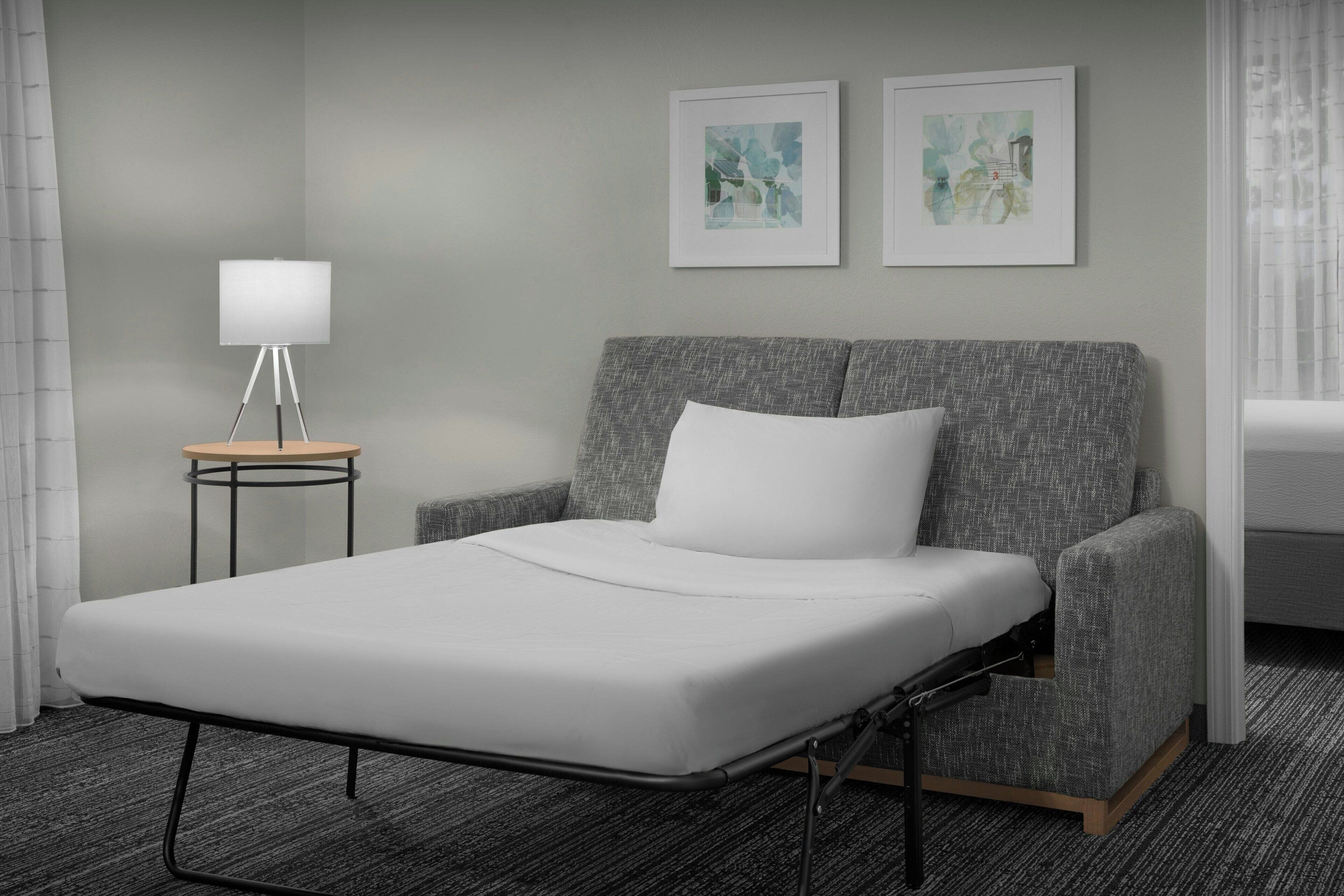 Une chambre d’hôtel moderne avec un lit simple, des fauteuils recouverts d’un tissu gris, une petite table avec une lampe et deux œuvres d’art encadrées accrochées au mur.