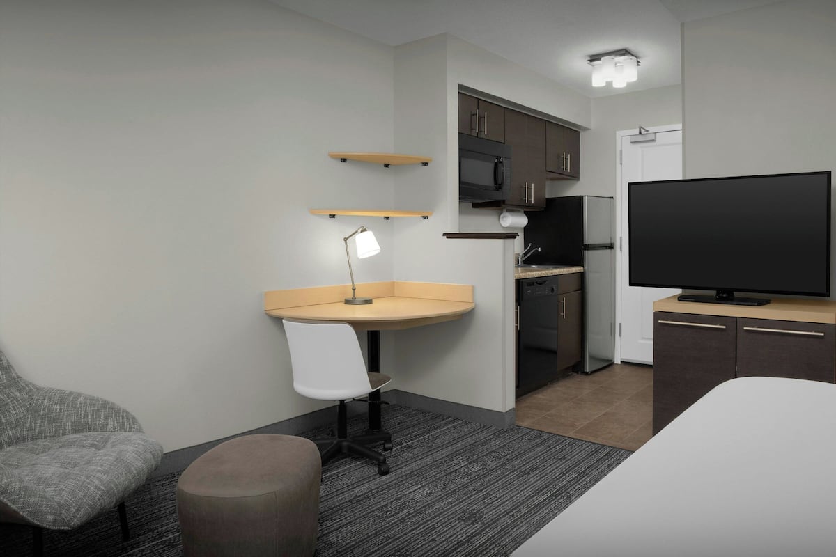 Ein kompaktes Hotelzimmer mit Küchenzeile, Schreibtisch mit Lampe, Fernseher, Sessel und Bett.