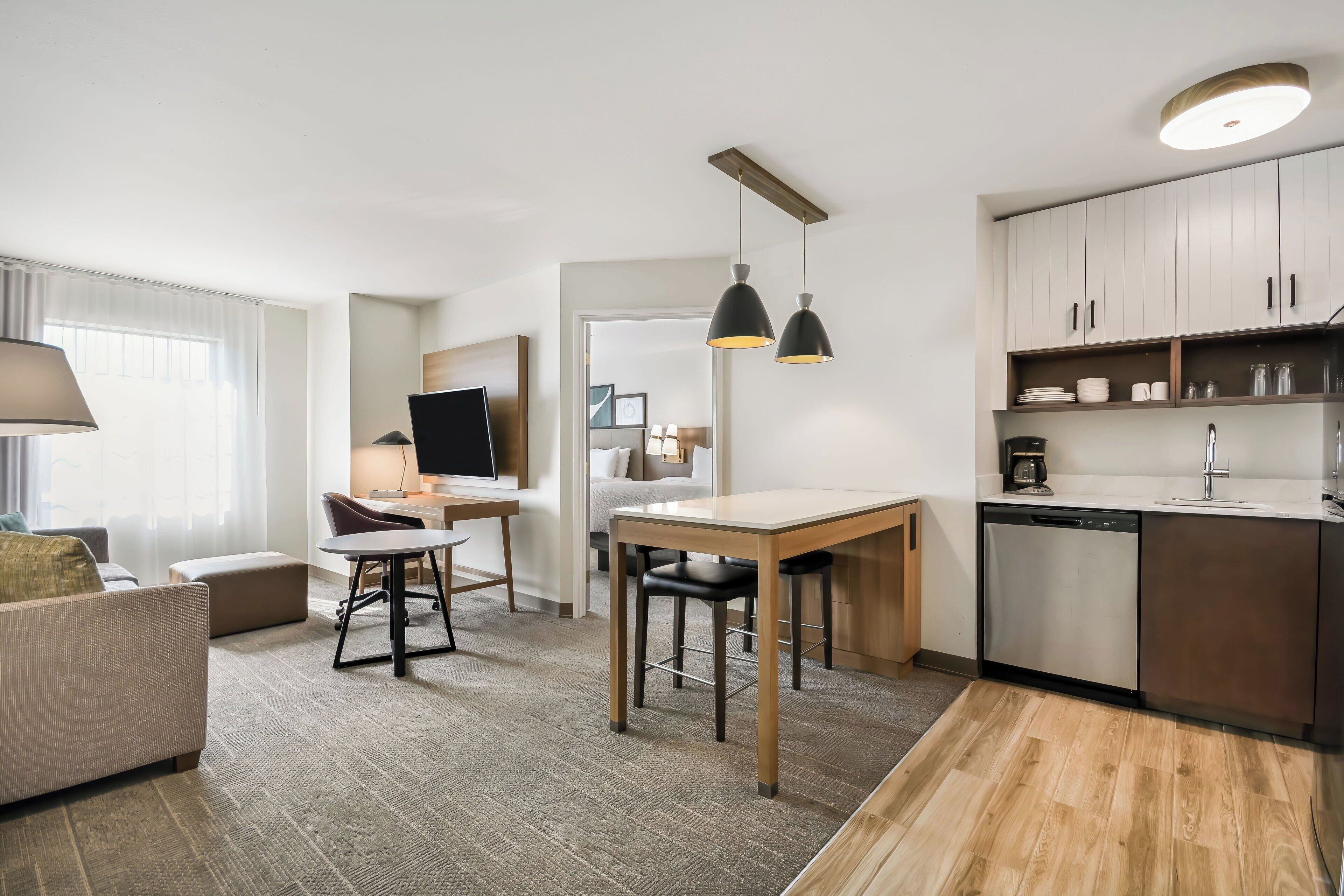 Habitación de hotel moderna con sofá, una mesa de comedor con sillas, televisor, zona de cocina con armarios y lavavajillas, y un ventanal con cortinas.