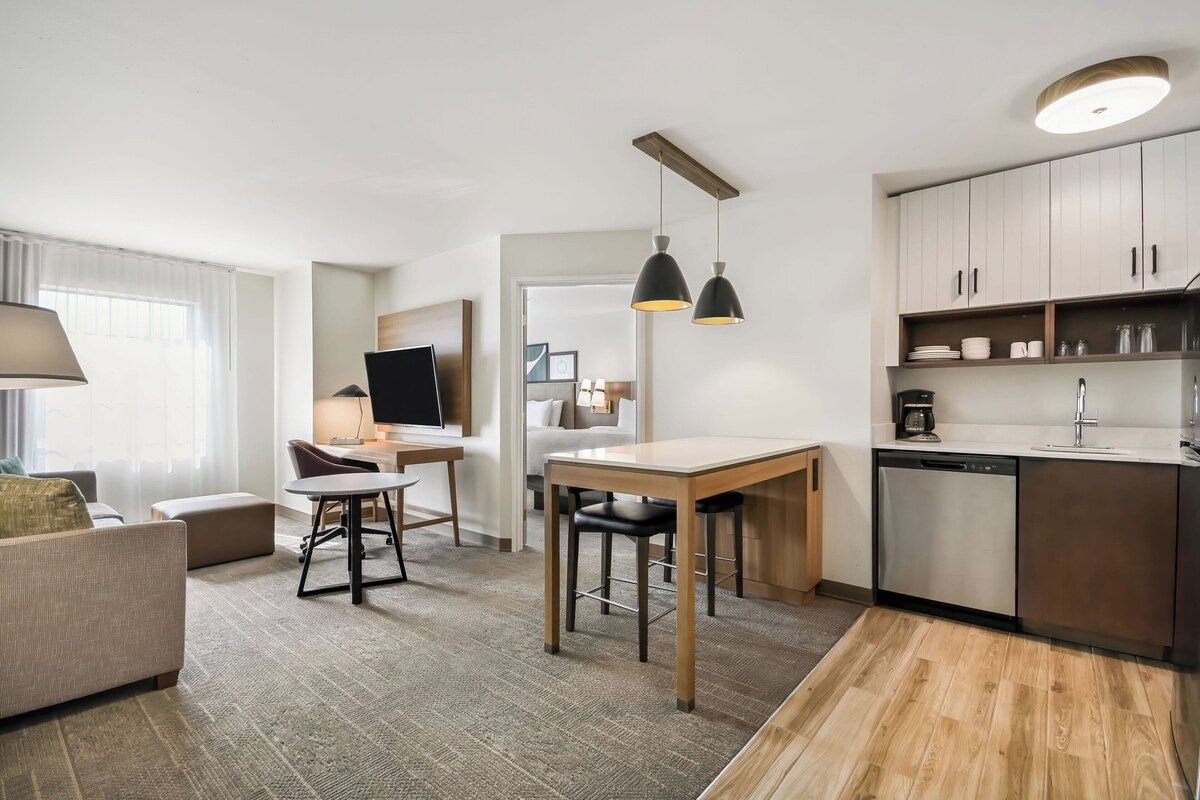 A modern hotel room with a sofa, a dining table with chairs, a TV, a kitchen area with cabinets and a dishwasher, and a large window with curtains.