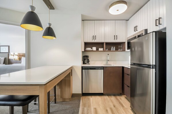 Una cocina moderna con un refrigerador de acero inoxidable, lavavajillas y cafetera. Hay un banco de madera en la encimera de la cocina y una mesa de comedor con banco. La cocina tiene gabinetes blancos y una encimera de color claro.