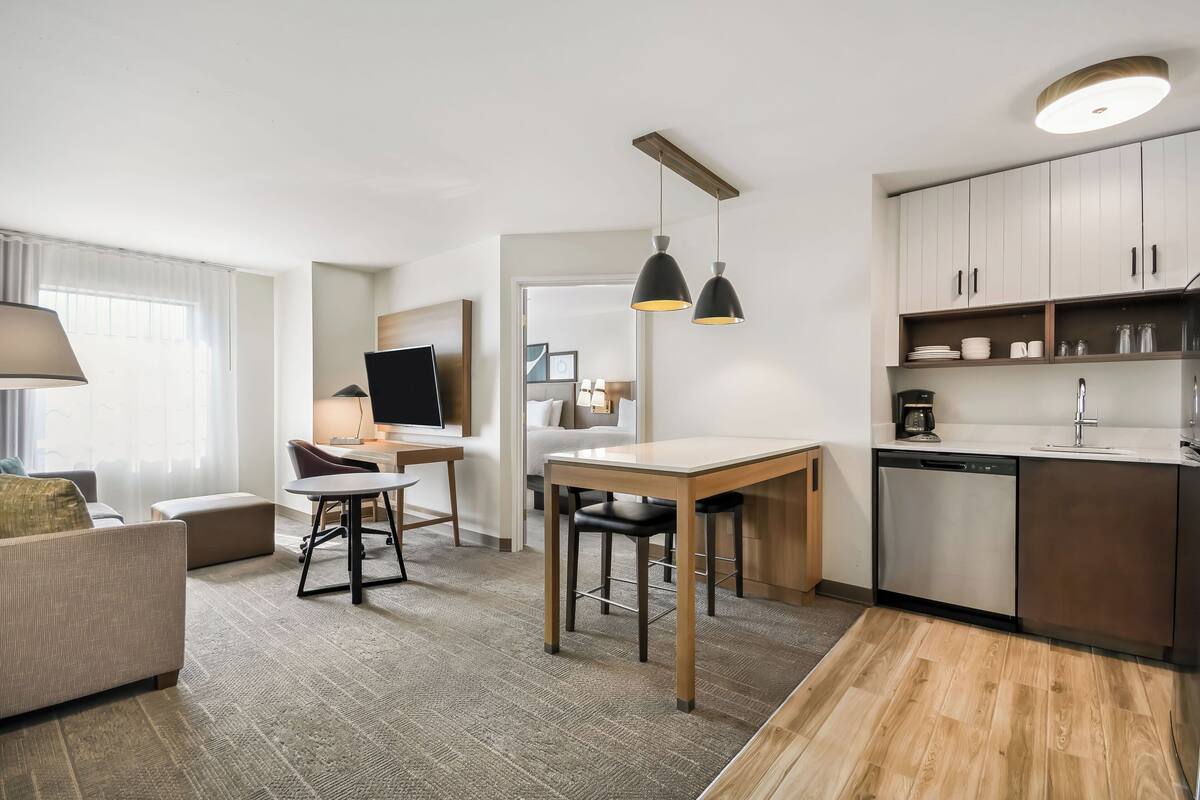 A modern hotel room with a sofa, a dining table with chairs, a TV, a kitchen area with cabinets and a dishwasher, and a large window with curtains.