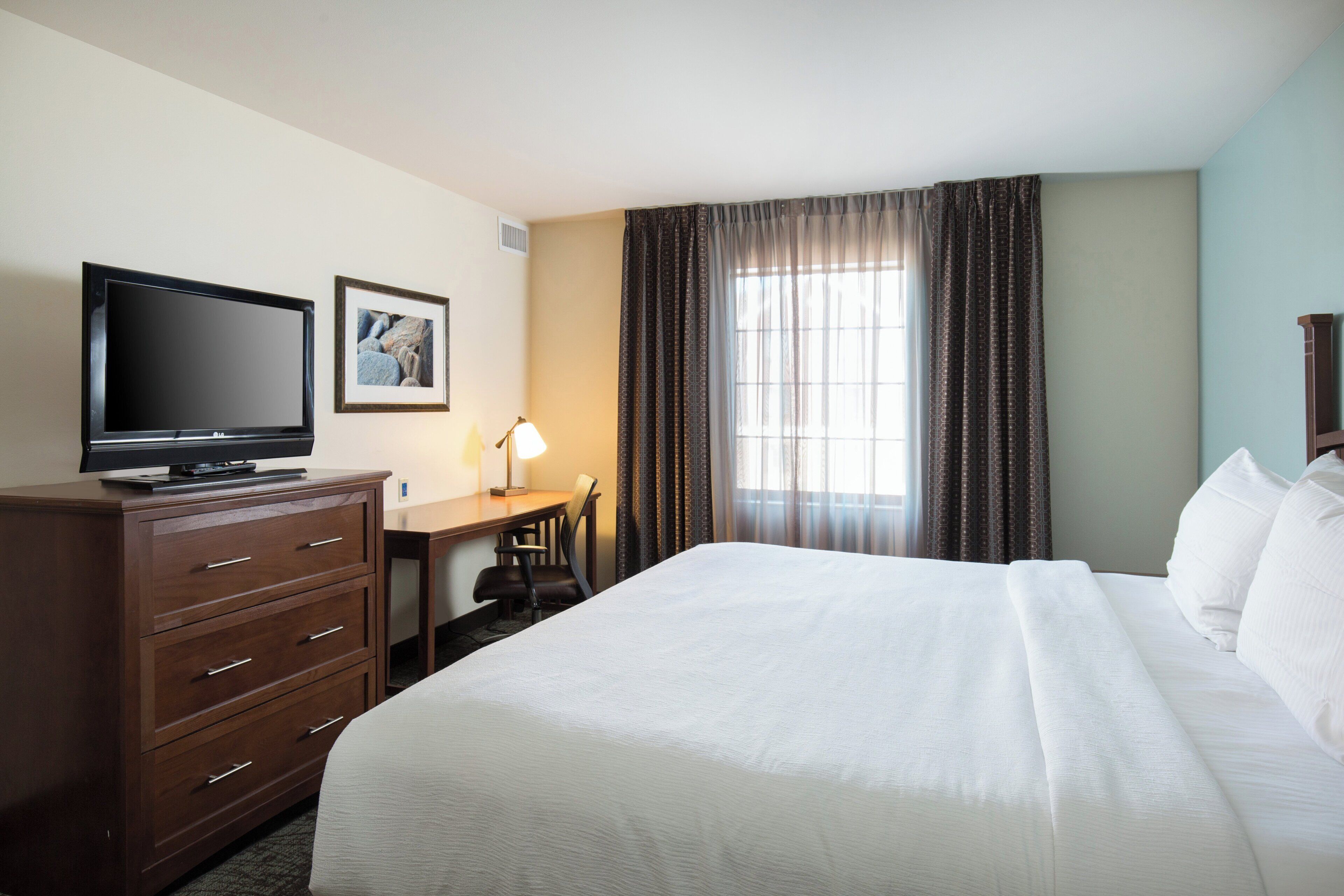 Habitación de hotel con una cama grande, un escritorio con una silla, un televisor y una ventana con cortinas.