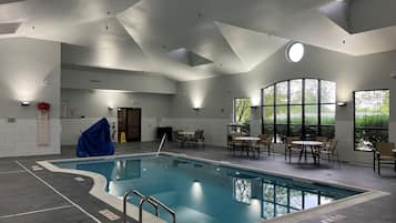 Une piscine dotée d’une terrasse carrelée, entourée de tables et de chaises, et de grandes fenêtres offrant une vue sur la verdure.