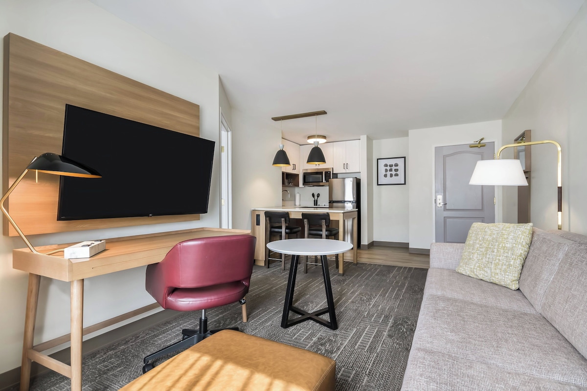 A modern hotel room with a flat-screen TV, a wooden desk, a red chair, a round table, a sofa, and a kitchenette in the background.