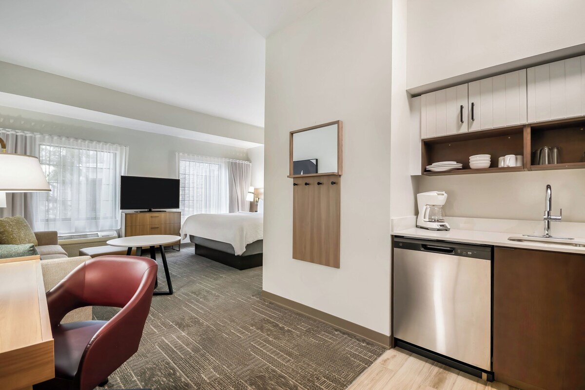A hotel room with a bed, a desk, a chair, a TV, a kitchen area with a sink and dishwasher, and a window with curtains.