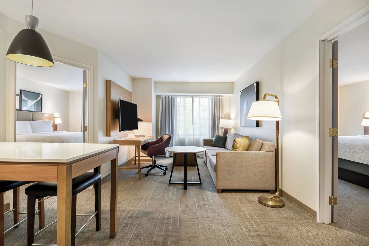 A hotel room with a dining area, a sofa, a TV, and a large window with curtains.