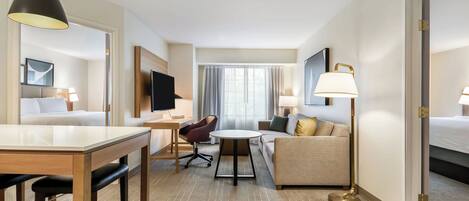 A hotel room with a dining area, a sofa, a TV, and a large window with curtains.