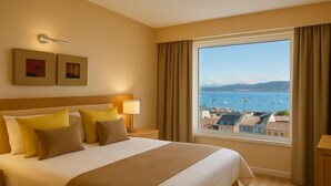 A hotel room with a large bed, two bedside lamps, a view of the sea and buildings, and a window with curtains.