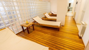 A spa room with wooden flooring, two sunbeds, a wooden table with a candle, and a frosted glass partition.
