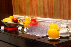 Un bufé de desayuno con un bol de fruta, una jarra de jugo de naranja y varios vasos vacíos sobre una encimera.