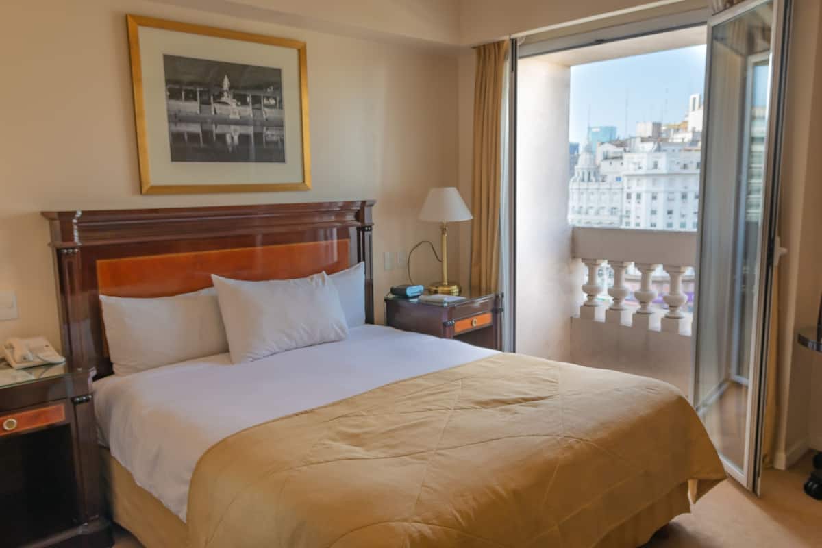 Une chambre d’hôtel avec un grand lit, une table de chevet avec une lampe, un tableau encadré au mur et une vue sur la ville par la fenêtre.