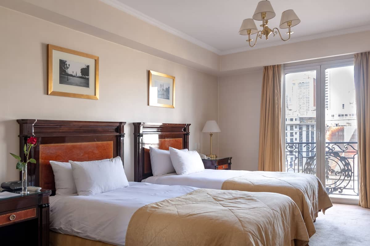 Une chambre d’hôtel avec deux lits, une tête de lit en bois, une table de chevet avec une lampe, un vase contenant une rose et une vue sur le paysage urbain par la fenêtre.