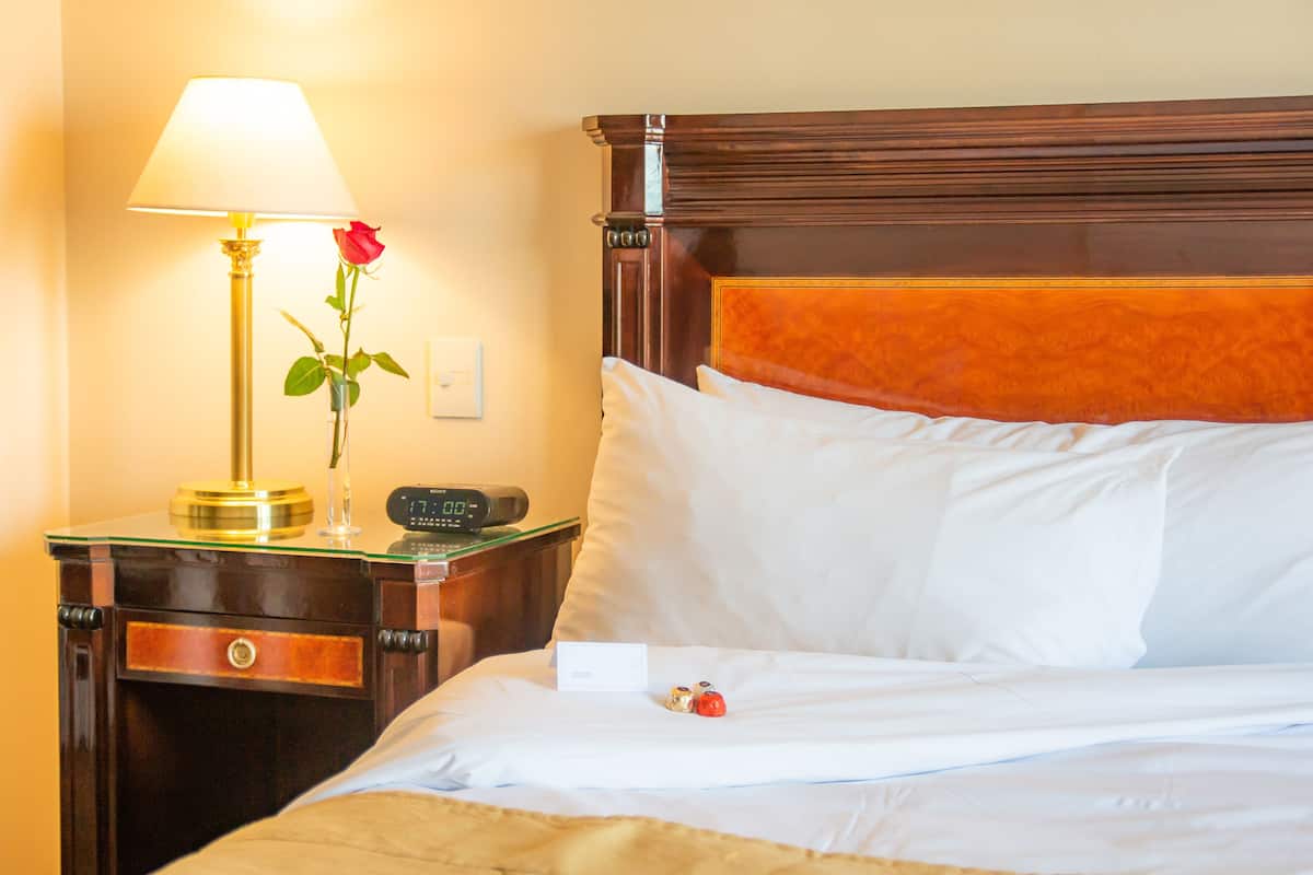 Une chambre d’hôtel avec une tête de lit en bois, une table de chevet avec une lampe, un vase contenant une seule rose et une horloge numérique.
