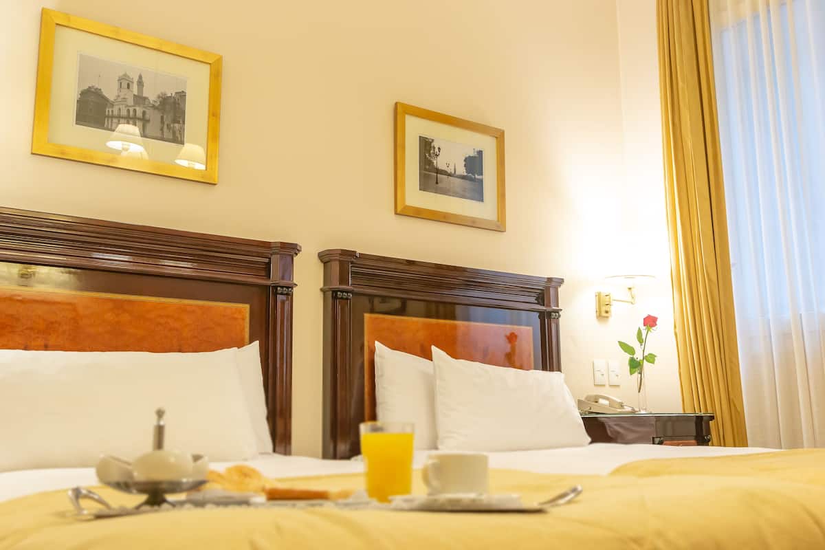 Une chambre d’hôtel avec deux lits, un plateau de petit-déjeuner, un vase contenant une rose rouge et des tableaux encadrés accrochés au mur.