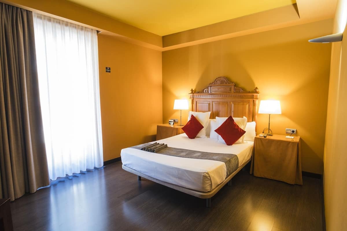 A hotel room with a large bed, two bedside tables, a wooden headboard, and a view of the outside through sheer curtains.