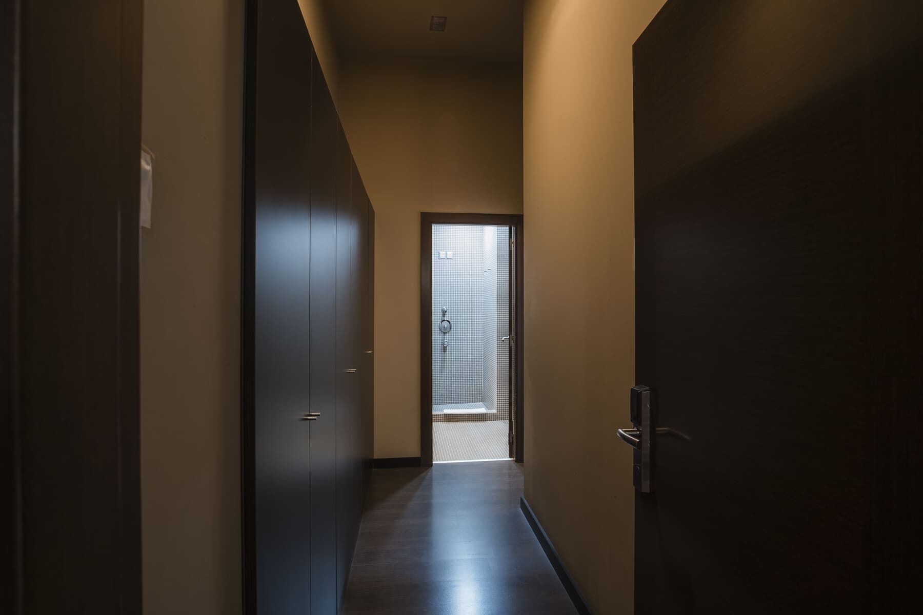 A narrow hallway with dark wooden doors on both sides, leading to a room with a large window.