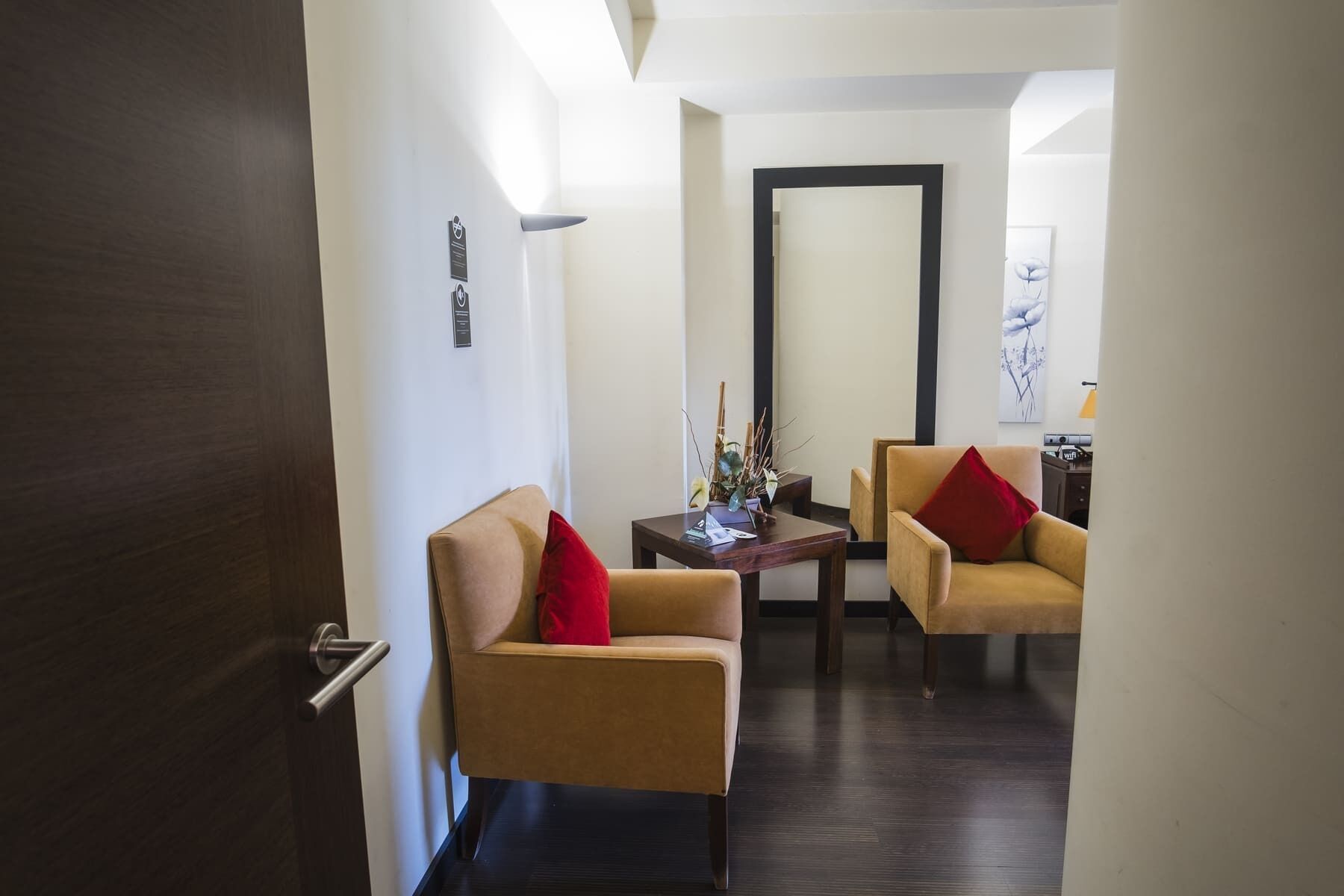 Une chambre d’hôtel avec une porte en bois, deux fauteuils beiges ornés de coussins rouges, une petite table en bois décorée de quelques objets, et un grand miroir accroché au mur.