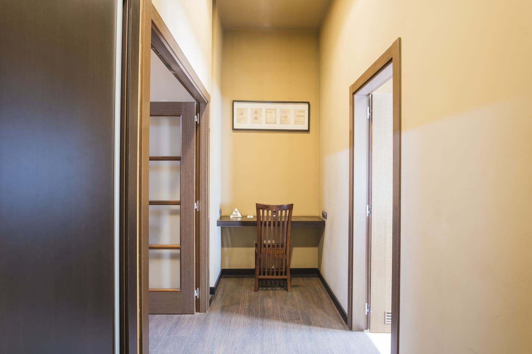 A narrow hallway with a wooden chair and a desk, a framed certificate on the wall, and a door on the left.