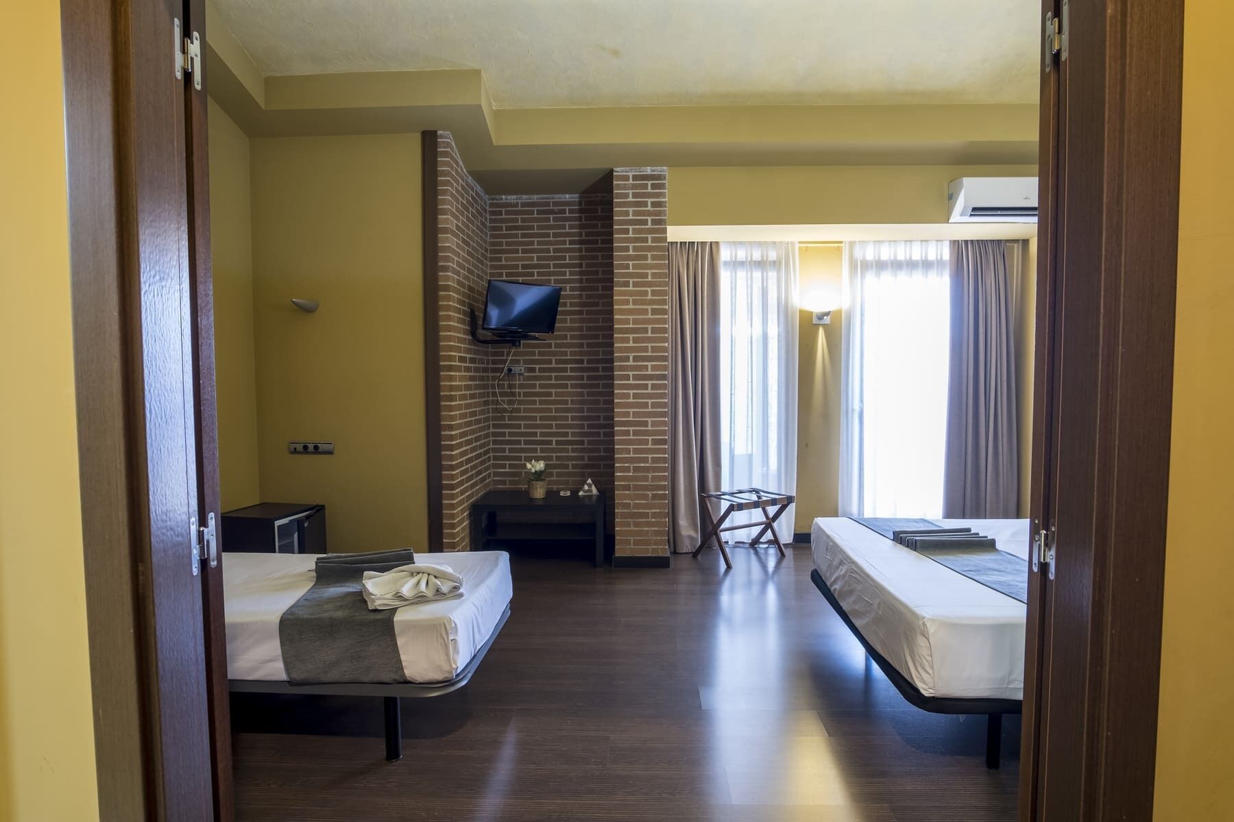 A hotel room with two beds, a TV mounted on a brick wall, a small table, and a window with curtains.