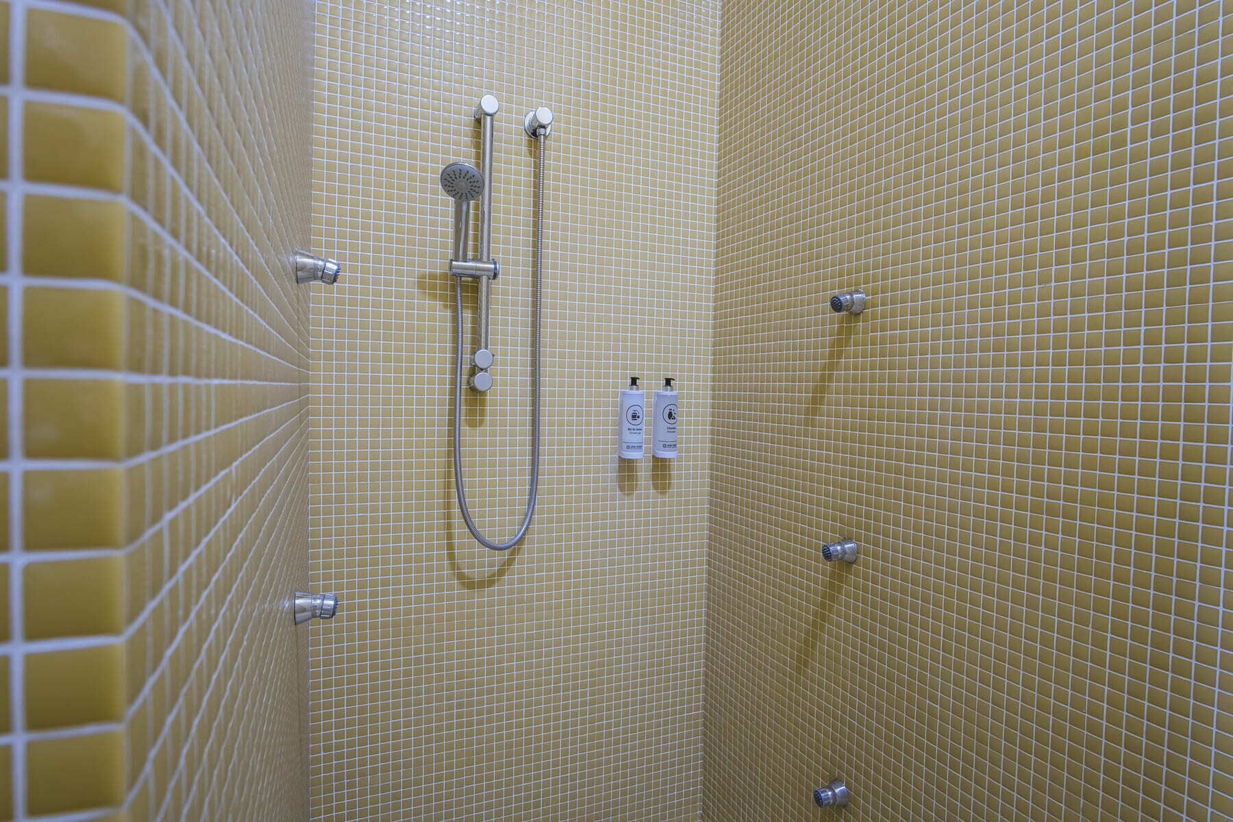 Ducha con cabezal de ducha de mano y grifo empotrado en la pared, paredes con azulejos y dispensador de toallas.