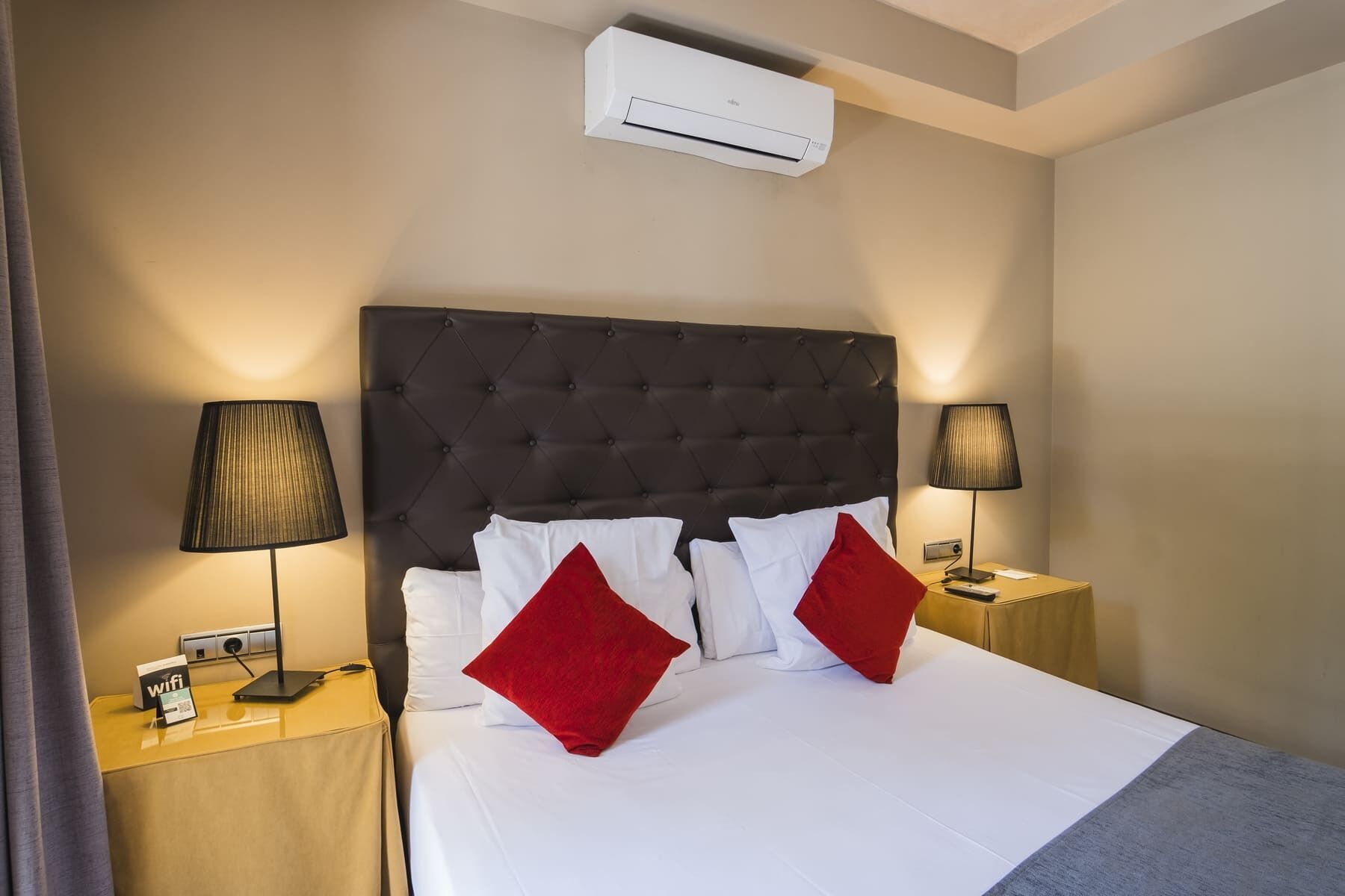 A hotel room with a large bed, two bedside lamps, a wall-mounted air conditioner, and a small table with a WiFi sign.