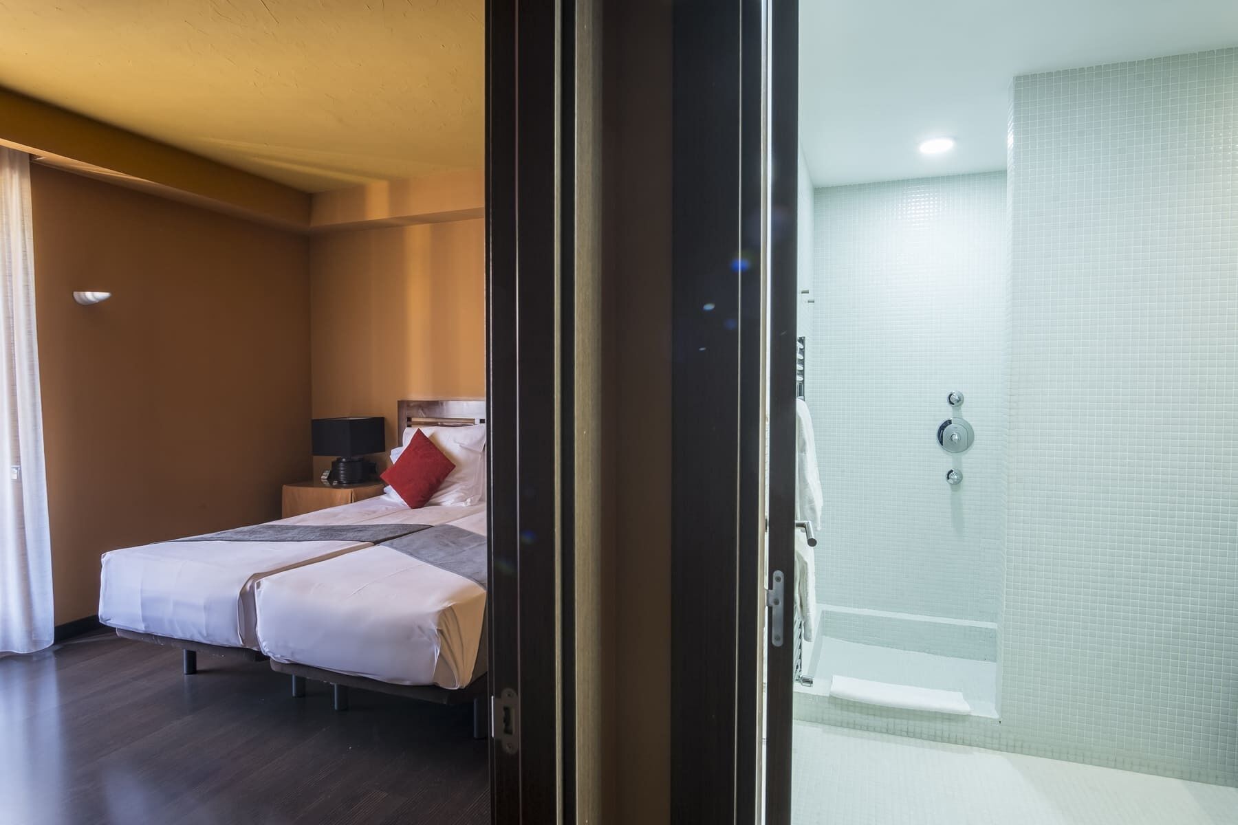 Een hotelkamer met een bed, nachtkastje, lamp en uitzicht op een badkamer met bad en douche.