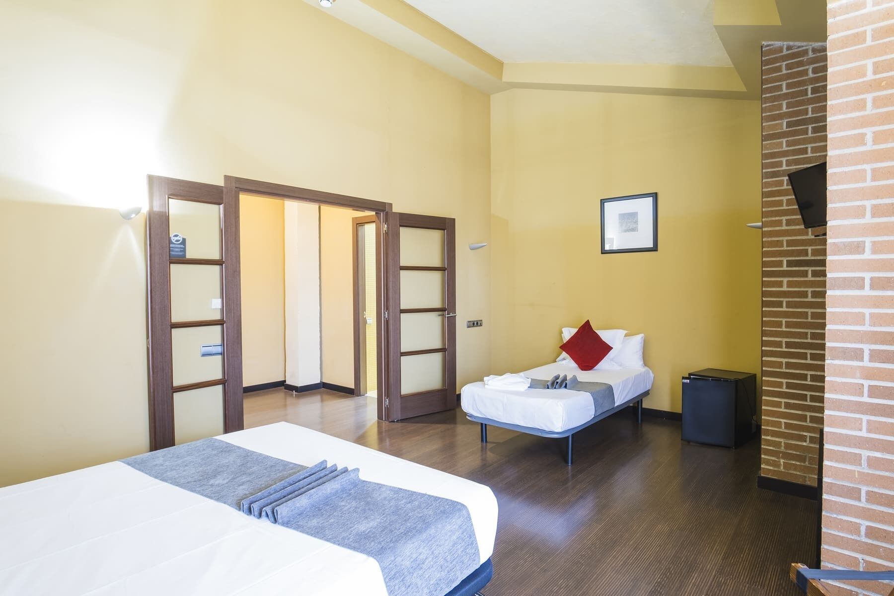 Una habitación de hotel con una cama, una mesita de noche pequeña, una silla y una puerta que da a otra estancia.