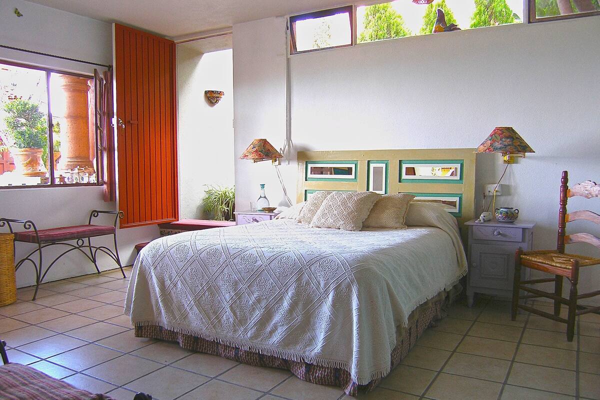 Une chambre à coucher comprenant un lit, des tables de chevet, une chaise, une fenêtre avec des rideaux et une vue sur la verdure à l’extérieur.