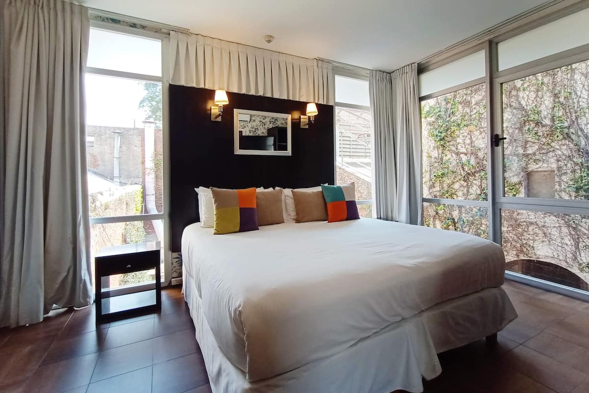 A bedroom with a large bed, a nightstand, a mirror, and two windows with curtains.