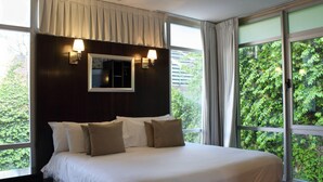 A bedroom with a large bed, two bedside lamps, a window with curtains, and a view of greenery outside. - Ribera Sur Hotel (Buenos Aires)