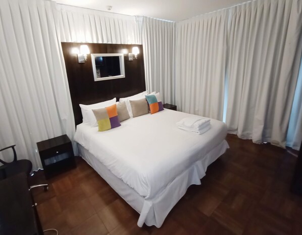 A hotel room with a large bed, white curtains, a flat-screen TV, and a wooden floor.
